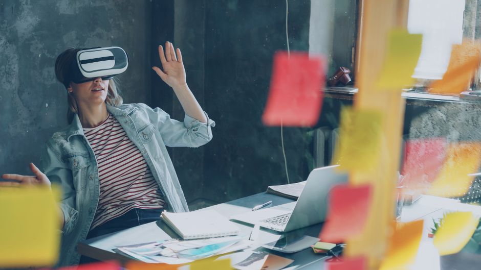 Mujer usando visor VR en escritorio con notas y laptop